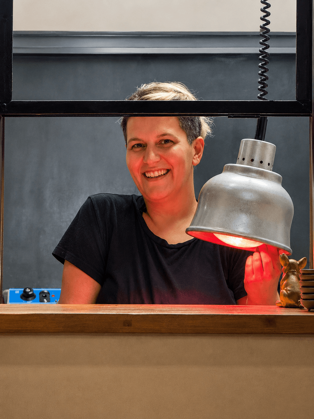 Donna sorridente in maglietta nera dietro un bancone con una lampada riscaldante a luce rossa.